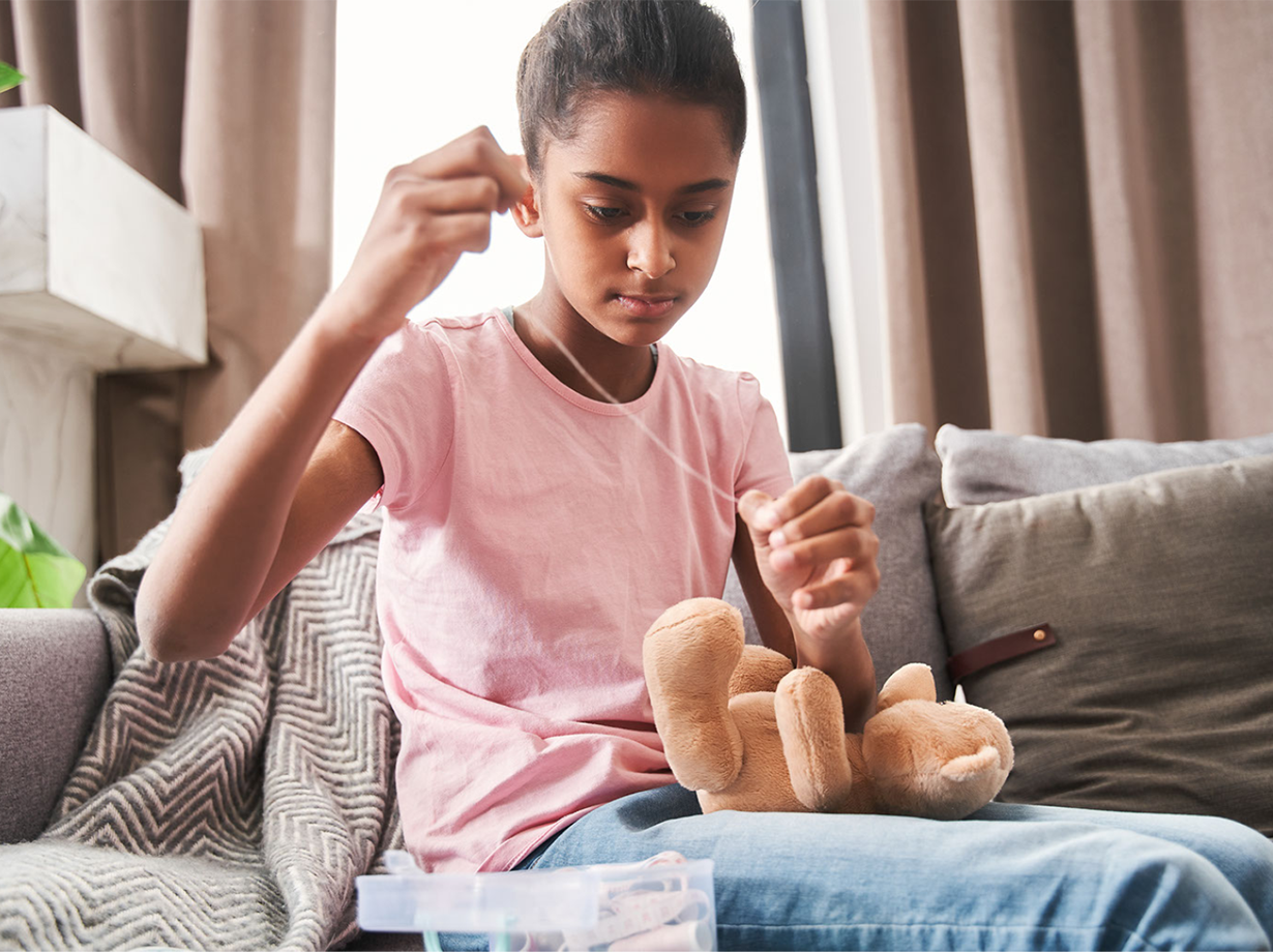 A child sewing a teadybear by hand while sitting comfortably on the couch